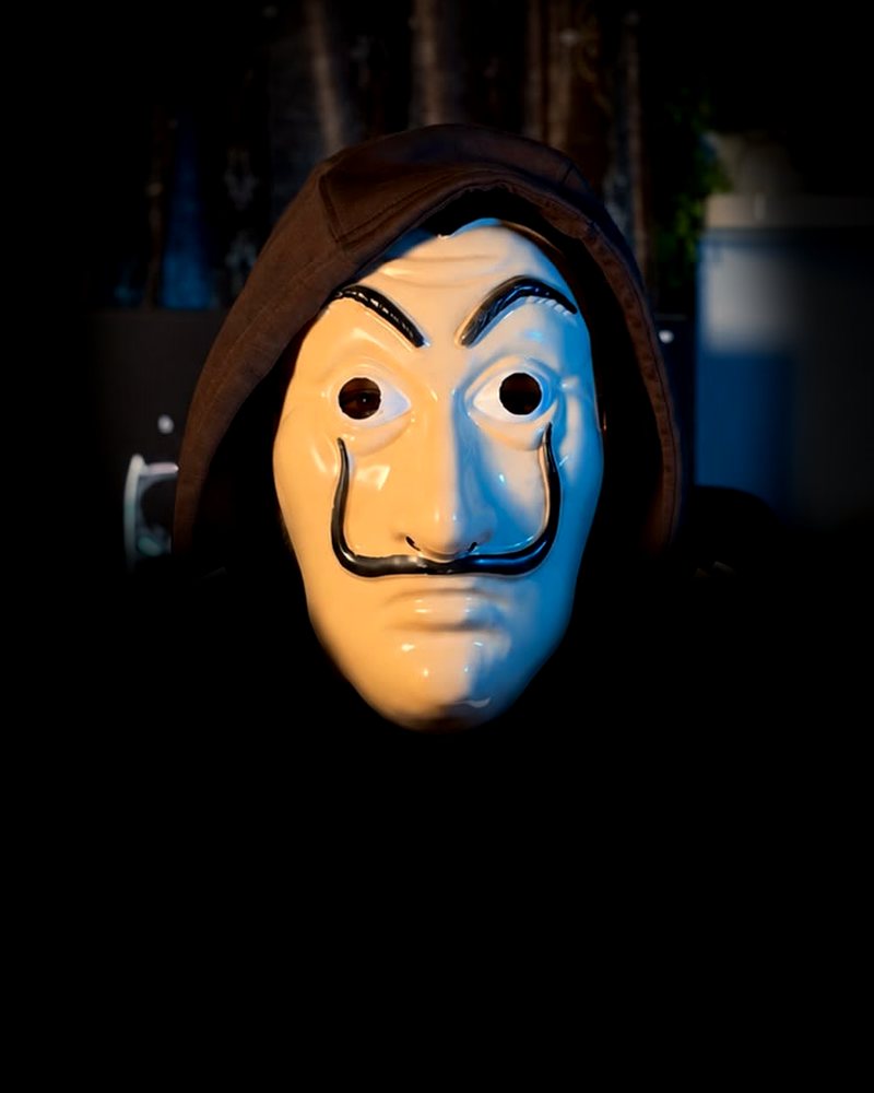 The Masked Mentor in a dark hood and a white theatrical mask